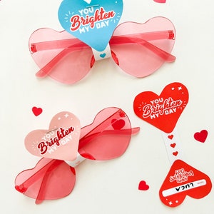 May include: Two pairs of heart-shaped sunglasses with red frames and pink lenses. Each pair has a heart-shaped tag attached with the text "You brighten my day" in red and white lettering.