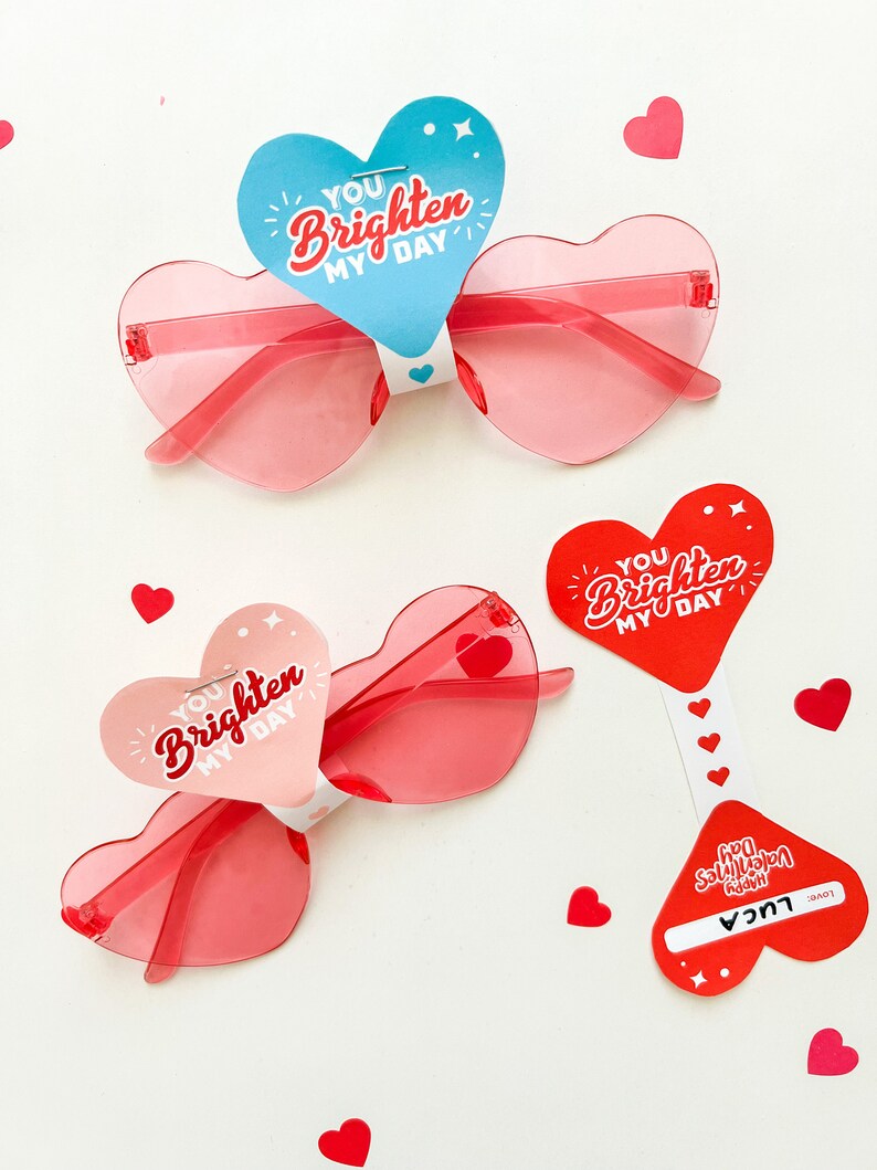 May include: Two pairs of heart-shaped sunglasses with red frames and pink lenses. Each pair has a heart-shaped tag attached with the text "You brighten my day" in red and white lettering.