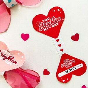 May include: Two heart-shaped Valentine's Day cards with the text "You brighten my day" and "Happy Valentine's Day" with a name and "Love" written on the bottom. The cards are next to pink heart-shaped sunglasses.