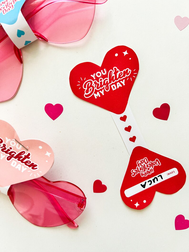 May include: Two heart-shaped Valentine's Day cards with the text "You brighten my day" and "Happy Valentine's Day" with a name and "Love" written on the bottom. The cards are next to pink heart-shaped sunglasses.