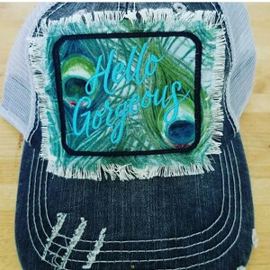 May include: A distressed denim trucker hat with a white mesh back. The front patch features a peacock feather design and the words "Hello Gorgeous" in turquoise script. The hat has a frayed edge.