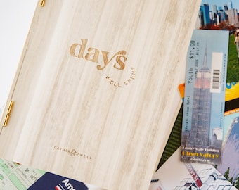 May include: A wooden box with the words "days well spent" and "gather & well" engraved on the front. The box is surrounded by various travel mementos, including a ticket to the Empire State Building, a seashell, and a photo of a person in New York City.