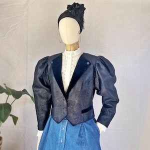 May include: A dark navy blue blazer with a subtle tonal swirl pattern, featuring structured puff sleeves, velvet lapels, and a single button closure. The blazer is worn over a white high-neck blouse with lace detailing and a blue denim skirt.
