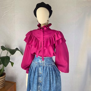 May include: A vibrant magenta long-sleeved blouse featuring a high ruffled collar, a cascading ruffled front panel, and long sleeves with gathered cuffs, paired with a high-waisted blue acid-wash denim skirt with a front button closure.