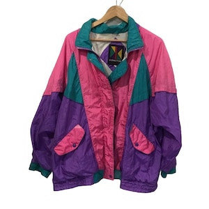 Puede incluir: Chaqueta vintage con bloques de color rosa, turquesa y morado. La chaqueta presenta un cuello alto, bolsillos delanteros y un parche con el logotipo. La chaqueta está colgada en una percha de madera.