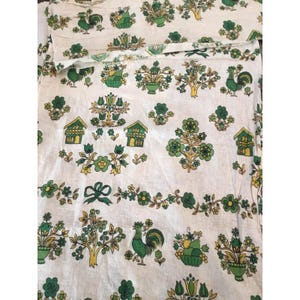 May include: A white fabric featuring a repeating pattern of green and yellow illustrations. The design includes roosters, small houses, potted plants, trees, and floral vines with bows, all rendered in a consistent color palette of green and yellow.