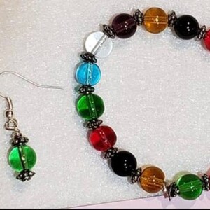 May include: A set of jewelry featuring a bracelet and earrings. The bracelet is made of colorful glass beads in shades of red, orange, yellow, green, blue, purple, and clear. The earrings are silver with green glass beads.