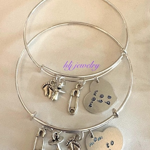 May include: Two silver bangle bracelets with charms. Charms include baby feet, pacifiers, safety pins, and heart-shaped pendants with the words "mom to be". The bracelets are on a white surface.