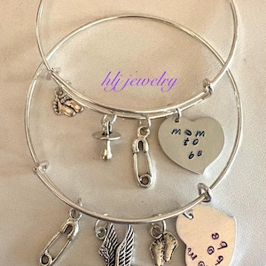 May include: Two silver-toned bangle bracelets with charms. Charms include a baby foot, pacifier, safety pin, heart with "mom to be" text, stork carrying a baby, and a heart with text. The bracelets are on a light-colored surface. The text "hlj jewelry" is visible.