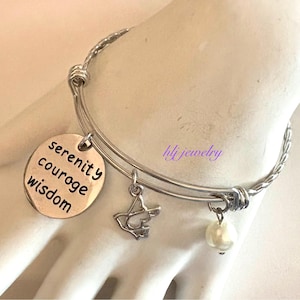 May include: Silver-toned bracelet with a braided band and three charms. One charm is a round pendant with the words "serenity courage wisdom". Other charms include a dove and a faux pearl. The bracelet is on a wrist.