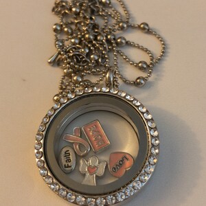 May include: A silver locket necklace with a clear glass insert and a silver chain. The locket contains several charms, including a pink ribbon, a heart, an angel, and charms with the words "Faith", "Hope", and "Love".