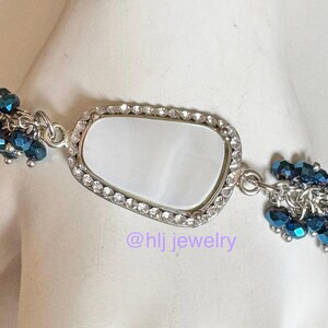 May include: Silver bracelet with a rectangular, white center stone, framed by clear crystals. Clusters of dark blue beads dangle from each side. The text "@hlj jewelry" is visible.