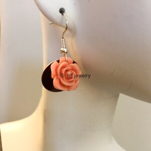 May include: A pair of dangle earrings with a peach-colored rose flower charm on a silver-toned circular base. The earrings are on a mannequin's ear. The text "© hlj jewelry" is visible in the image.