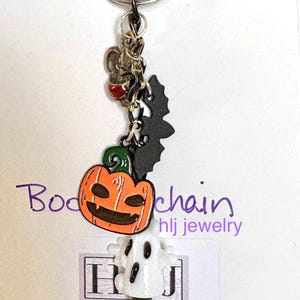 May include: A Halloween-themed keychain featuring a smiling orange pumpkin, a black bat, and a white ghost charm. The keychain includes a metal ring and a small charm with a red accent. The text "Body chain hlj jewelry" is visible.