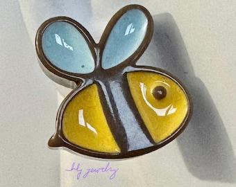 Könnte beinhalten: Ein Keramik-Bienen-Anhänger mit gelben und braunen Streifen, blauen Flügeln und einem braunen Auge. Der Anhänger befindet sich auf einem weißen Hintergrund.
