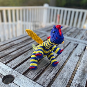 May include: A blue and yellow striped crocheted dragon with red accents and yellow wings. The dragon is sitting on a wooden table.