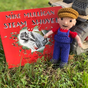 May include: A crocheted doll wearing a brown cap, red shirt, and blue overalls stands in front of a red book titled "Mike Mulligan and His Steam Shovel".