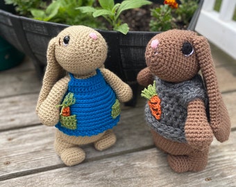 May include: Two crocheted bunny toys, one brown and one beige, are sitting on a wooden surface. The brown bunny is wearing a gray sweater with a carrot applique. The beige bunny is wearing a blue overall with green pockets and carrot appliques.