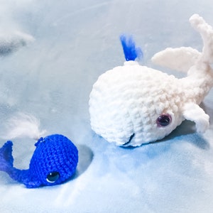 Puede incluir: Dos ballenas de ganchillo hechas a mano, una blanca y otra azul, descansan sobre una superficie azul claro. La ballena blanca tiene una cola curvada y un mechón azul. La ballena azul tiene una cola curvada y un mechón blanco. Ambas tienen ojos oscuros.