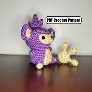 May include: A crocheted purple and yellow amigurumi toy resembling a monkey with a long tail. The toy is sitting on a wooden surface. PDF Crochet Pattern.