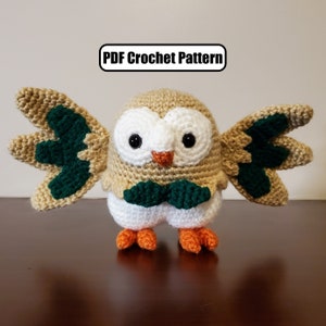 May include: Crocheted owl amigurumi toy with brown, white, and green yarn. The owl has large eyes and a small orange beak. The toy is standing on a brown surface. PDF Crochet Pattern.