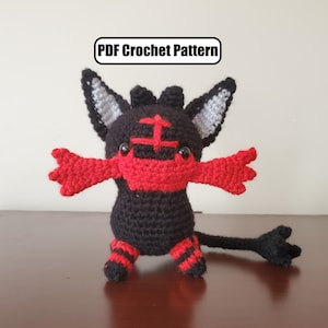 May include: Crocheted plush toy of a black and red Pokemon character with a long tail. The toy has a red mouth and black eyes. The text "PDF Crochet Pattern" is visible above the toy.