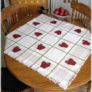 May include: A white table runner with a red apple design. The runner has a fringed edge and is laid on a wooden table.