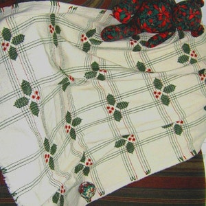 May include: Cream-coloured plaid throw blanket with green holly and red berry accents. A teddy bear with a poinsettia pattern sits to the left of the blanket. A decorative ball with a similar pattern is on the right.