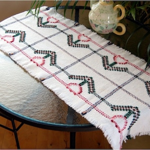 May include: White table runner with a woven design featuring pink, green and blue arrows. The runner has a fringed edge and is displayed on a glass table.
