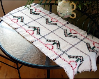 May include: White table runner with a woven design featuring pink, green, and blue arrows. The runner has a fringed edge and is displayed on a glass table.