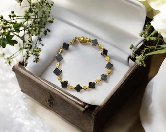 Puede incluir: Un brazalete dorado con abalorios negros en forma de trébol se exhibe dentro de una caja de madera. El brazalete está hecho de metal dorado y presenta un cierre magnético. La caja está forrada con tela blanca y rodeada de vegetación.