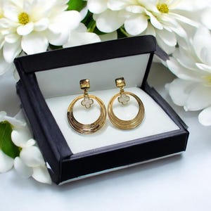 Peut inclure: Une paire de boucles d'oreilles dorées en forme de cerceau avec un charm en forme de trèfle, présentée dans un coffret noir avec un intérieur blanc. Les boucles d'oreilles ont un design à plusieurs niveaux, sur fond de fleurs blanches.