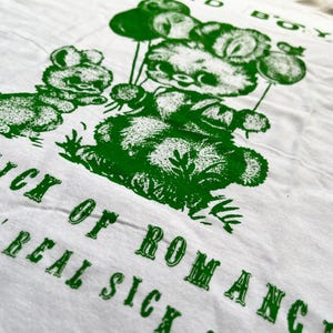 May include: White t-shirt with a green graphic of cartoon bears holding balloons. The text "BAD BOY" is at the top, and "SICK OF ROMANCE, REAL SICK" is printed at the bottom in a vintage font.