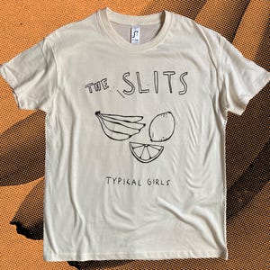 Könnte beinhalten: Ein weißes T-Shirt mit einem schwarzen grafischen Aufdruck der Band "The Slits" mit einem Bananen-, Zitronen- und Limetten-Design. Der Text "Typical Girls" ist unter dem Bandnamen gedruckt.