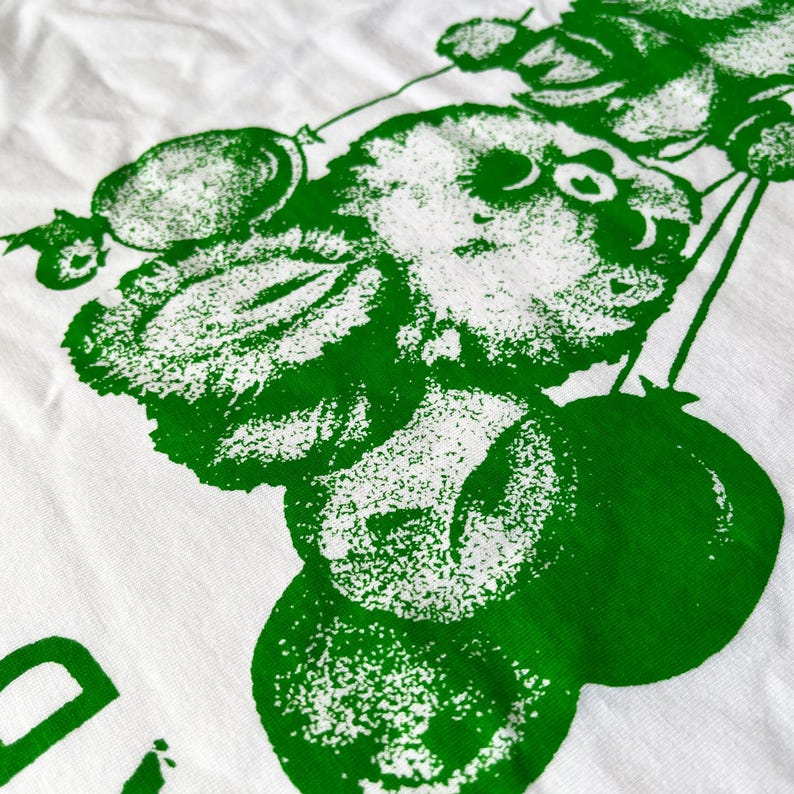 May include: White t-shirt with a large green graphic print. The design features a stylized illustration of a cat holding balloons and fruit. The print has a textured, speckled appearance.