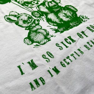 May include: White t-shirt with a green graphic of teddy bears holding balloons. The text "I'M SO SICK OF IT AND I'M GETTIN' REAL" is printed in green below the bears. The shirt is made of a soft, comfortable material.