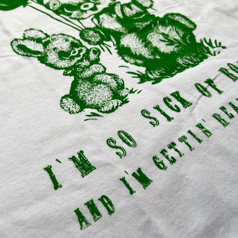 May include: White t-shirt with a green graphic of teddy bears holding balloons. The text "I'M SO SICK OF IT AND I'M GETTIN' REAL" is printed in green below the bears. The shirt is made of a soft, comfortable material.