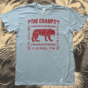 May include: Pale blue t-shirt with a red tiger graphic and the words "THE CRAMPS", "THE CITY IS A JUNGLE AND I'M A BEAST". The design includes palm trees, skulls, and a dripping pattern. The shirt is made of a soft material.