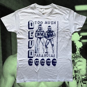 Op de afbeelding: Wit T-shirt met een blauwe print van twee worstelaars met maskers en de tekst "DEVO TOO MUCH PARANOIAS".