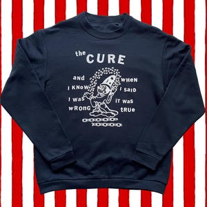 Op de afbeelding: Marineblauw sweatshirt met witte tekst en illustratie. De tekst luidt "the CURE" boven een stripfiguur met een tovenaars hoed. Daaronder staan de woorden "and I KNOW I was WRONG when I said it was tRUE".