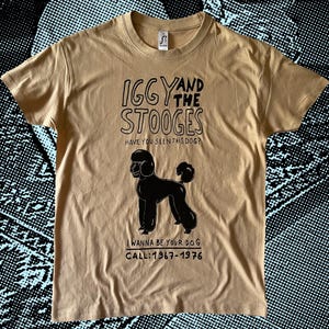 以下が含まれることがあります： 黒い文字と黒いプードルのイラストが描かれたタン色のTシャツ。「Iggy and the Stooges」と「Have you seen this dog?」の文字と、「I wanna be your dog」と「Call: 1967-1976」の文字がプードルの下にあります。
