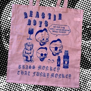 Peut inclure: Un sac fourre-tout rose clair avec une impression graphique bleu foncé. Le motif comprend le texte Beastie Boys, un singe, trois têtes de canard, un verre, une bouteille et une bulle avec le texte Me and the crew, we're drinking Brass Monkey.