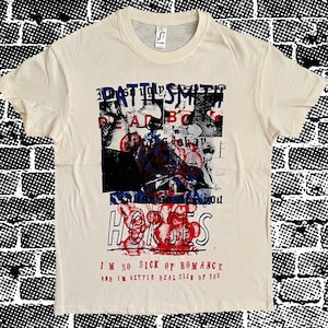 Op de afbeelding: Crèmekleurig T-shirt met een grafisch ontwerp met de tekst "PATTI SMITH" en "HORSES" in rood en blauw. Het shirt bevat ook de zinnen "I'M SO SICK OF ROMANCE" en "AND I'M GETTIN' REAL SICK OF YOU".