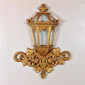 May include: A gold-colored wall-mounted lantern decoration featuring an open frame design. The base is adorned with intricate relief carvings of grape clusters and vine leaves, extending outward in a symmetrical scrollwork pattern.