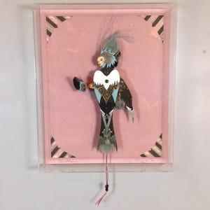 May include: A framed mixed media artwork featuring a colourful bird-like figure with a feathered headdress. The figure is made of paper, feathers, and other materials. The background is pink with black and white stripes.