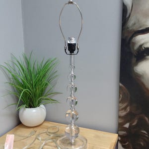 May include: A clear glass table lamp with a spherical base and a series of clear glass spheres stacked along the stem. The lamp has a silver-colored metal shade holder and a clear glass bulb finial. The lamp is on a wooden table.