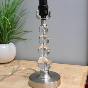 May include: A clear glass and brushed nickel table lamp with a black socket. The lamp features a base and a stem composed of four clear glass spheres. The lamp is on a wooden surface.
