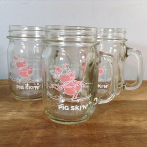 May include: Three clear glass mason jar mugs with handles. Each mug features a pink cartoon pig graphic and the text "PIG SKI'N" in white. The mugs are on a wooden surface.