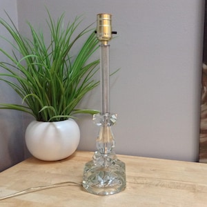 May include: A clear glass table lamp with a faceted base and a crystal-like design. The lamp has a tall, slender stem and a gold-coloured bulb socket. A green plant in a white pot is in the background.