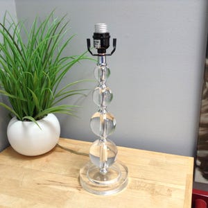 May include: A clear glass table lamp with a chrome base and accents. The lamp features three large, spherical glass orbs stacked on a chrome rod. The lamp sits on a round base and has a black lamp socket. A potted green plant is to the left.